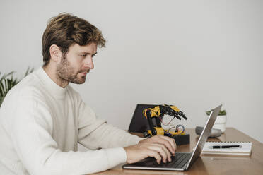 Businessman typing on laptop by robotic arm at workplace - EBBF06752