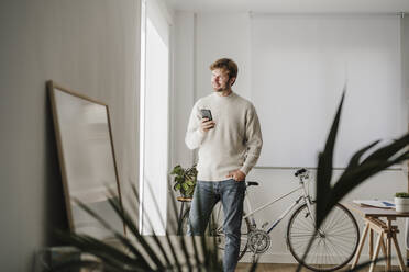 Geschäftsmann mit Smartphone schaut aus dem Fenster im Büro - EBBF06766