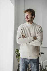 Happy businessman with arms crossed looking out of window in office - EBBF06768