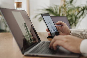 Hands of businessman using laptop and smart phone examining wind turbine model at office - EBBF06789