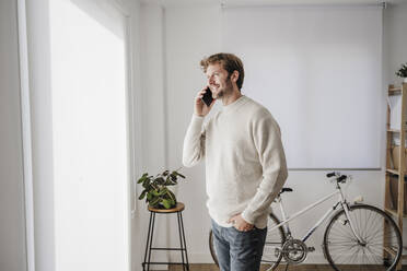 Happy businessman talking on smart phone and looking out of window at workplace - EBBF06804