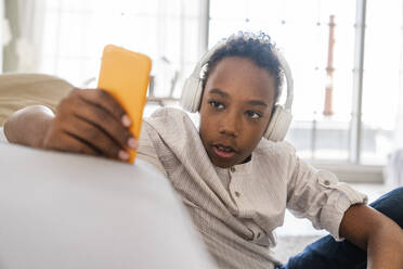 Junge, der ein Mobiltelefon benutzt und Kopfhörer trägt, während er zu Hause Musik hört - VPIF07656