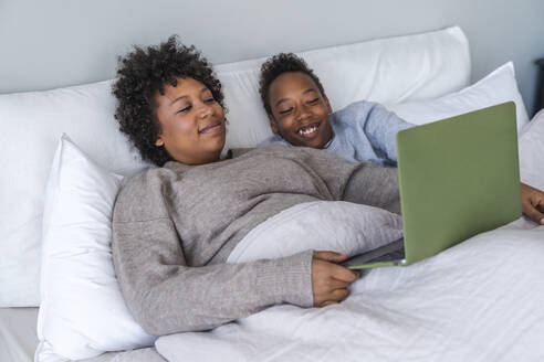 Happy son and mother watching movie using laptop in bedroom - VPIF07665