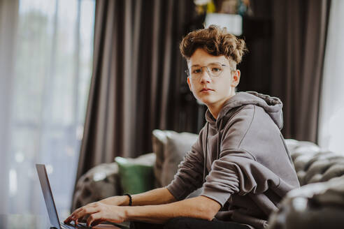 Teenage boy with laptop sitting on sofa at home - MDOF00166
