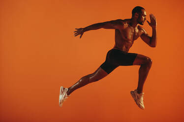 Athlete taking long stride forward during sprint. Bare chested athlete running hard on orange background. - JLPSF19425