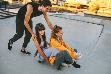 Glücklicher junger Mann, der seine Freunde auf dem Skateboard im Skatepark schiebt. Gruppe von Freunden, die sich im Skatepark vergnügen. - JLPSF19440