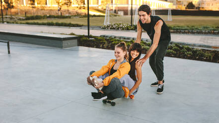 Zwei Mädchen sitzen auf einem Skateboard, das von einem männlichen Freund im Skatepark geschoben wird. Eine Gruppe von Freunden hat eine tolle Zeit im Skatepark. - JLPSF19441