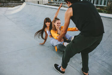 Zwei Mädchen sitzen auf einem Skateboard, das von einem männlichen Freund im Skatepark gezogen wird. Eine Gruppe von Freunden hängt im Skatepark ab. - JLPSF19442