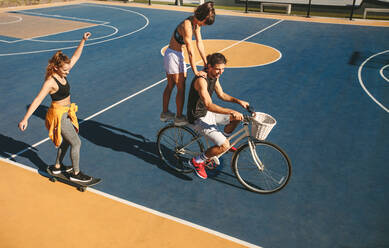 Ein Paar hat Spaß beim Fahrradfahren mit einer Freundin, die auf einem Basketballplatz im Freien Skateboard fährt. - JLPSF19466