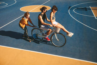 Eine Gruppe von Freunden hat Spaß beim Fahrrad- und Skateboardfahren auf einem Basketballplatz im Freien. Ein Mann mit zwei Freundinnen amüsiert sich im Freien. - JLPSF19467