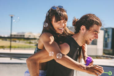 Couple having fun outdoors. Man piggybacking woman playing with soap bubble outdoors. - JLPSF19479