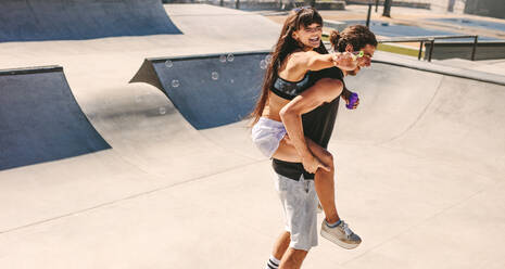 Man giving his boyfriend a piggyback whilst playing with soap bubbles at skate park. Young man and woman having a great time outdoors on a summer day. - JLPSF19481