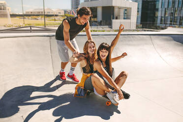Glücklicher junger Mann schiebt zwei Mädchen, die auf einem Skateboard in einem Skatepark sitzen. - JLPSF19490