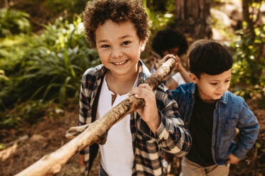 Niedlicher Junge trägt einen Stock mit Freunden im Wald. Kinder spielen ein Abenteuerspiel im Wald. - JLPSF19495