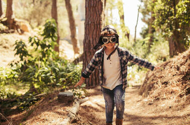 Kind mit Pilotenbrille und Hut, das im Freien im Wald läuft und so tut, als wäre es ein Pilot, der im Wald fliegt. - JLPSF19516