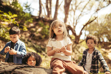Nettes Mädchen sitzt auf einem Baumstamm mit Stöcken in der Hand und ihre Freunde im Wald. Jungen und ein Mädchen spielen im Wald. - JLPSF19517