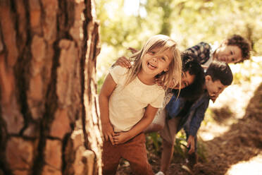 Eine Gruppe von Kindern steht in einer Reihe und späht hinter einem Baum im Freien. Kinder spielen Verstecken in einem Park. - JLPSF19518