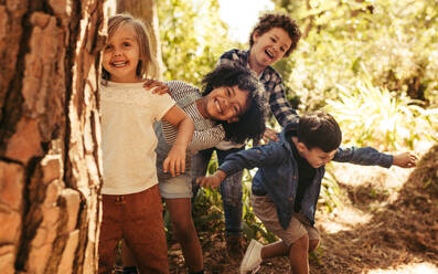 Niedliche, lächelnde Kinder, die hinter einem Baum im Park hervorschauen. Eine Gruppe von Kindern genießt es, in einem Wald Verstecken zu spielen. - JLPSF19519