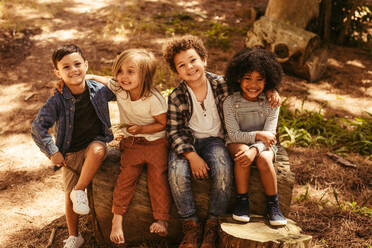 Eine Gruppe von vier Kindern sitzt auf einem Holzklotz im Freien. Eine multiethnische Gruppe von Kindern spielt zusammen in einem Wald. - JLPSF19520
