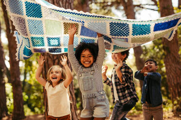 Vier Kinder im Park tanzen unter einer Picknickdecke. Gruppe von Kindern, die zusammen im Park Spaß haben. - JLPSF19535