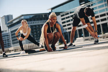 Group of people during workout session outdoors. Friends stretching on exercise mat outdoors in the city. - JLPSF19576