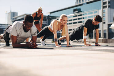 Eine Gruppe von Menschen, die im Freien auf einer Fitnessmatte ihre Grundübungen machen. Eine sportliche Gruppe trainiert gemeinsam in der Stadt. - JLPSF19583