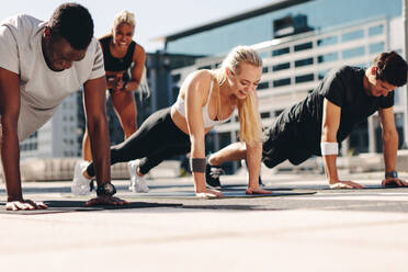 Fitness men and women doing push-ups with motivation from their female trainer outdoors in the city. - JLPSF19584