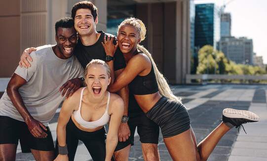 Fitte junge Freunde stehen und lachen in die Kamera, nachdem sie im Freien trainiert haben. Gruppe von vier jungen Leuten in Sportkleidung nach einer Trainingseinheit in der Stadt. - JLPSF19590