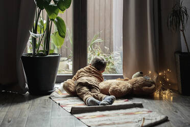 Junge schaut aus dem Fenster und liegt mit Stofftier zu Hause auf dem Teppich - VSNF00062