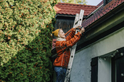 Smiling woman standing on ladder decorating house with lights for Christmas - VSNF00065