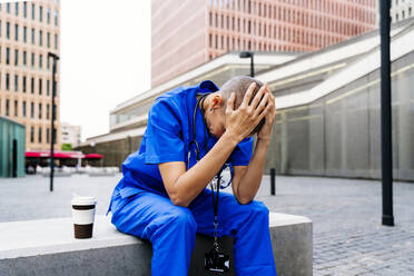 Ältere weibliche Krankenschwester sitzt mit dem Kopf in den Händen auf einer Bank - OIPF02543