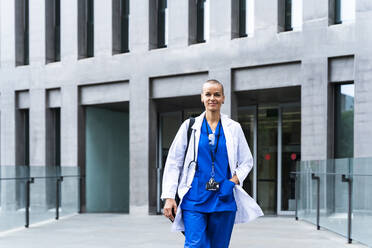 Lächelnde Ärztin mit Hand in der Tasche vor einem Krankenhaus - OIPF02581