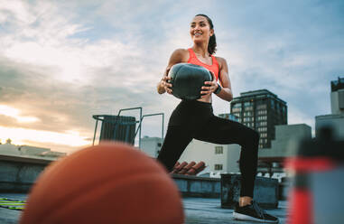 Female athlete doing forward lunges holding a medicine ball on rooftop. Fitness woman doing workout using medicine ball with a basketball and water bottle in the foreground. - JLPSF19653