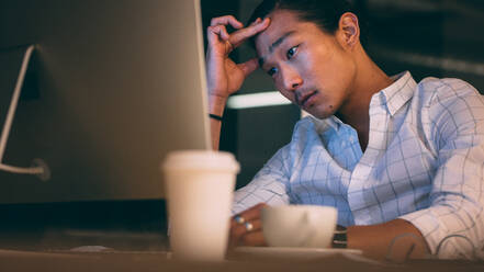 Unternehmer, der bis spät in die Nacht im Büro arbeitet und gestresst aussieht. Asiatischer Geschäftsmann, der mit der Hand auf der Stirn auf den Computerbildschirm schaut und nach einer Lösung sucht. - JLPSF19656