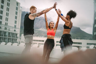 Drei Fitness-Leute stehen nach dem Training auf einem Dach und halten sich freudig an den Händen. Sportler geben sich an einem bewölkten Morgen auf der Terrasse gegenseitig High Five. - JLPSF19658