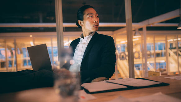 Asiatischer Geschäftsmann sitzt spät abends im Büro und arbeitet am Laptop. Unternehmer schaut zurück, während er spät im Büro arbeitet. - JLPSF19662