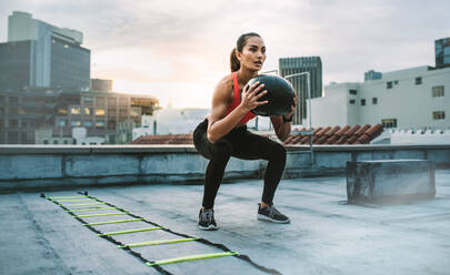 Weibliche Athletin macht Kniebeugen mit einem Medizinball auf einem Dach stehend. Frau macht Training mit Medizinball mit einer Agility-Leiter an ihrer Seite auf einem Dach. - JLPSF19667