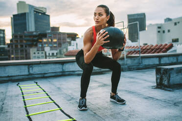 Fitness-Frau macht Training auf dem Dach mit einem Medizinball. Frau macht Kniebeugen mit einem Medizinball mit einer Agility-Leiter an ihrer Seite auf dem Dach. - JLPSF19668