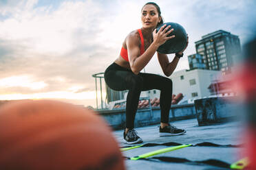 Niedriger Blickwinkel auf eine Frau, die Fitnessübungen auf der Terrasse eines Gebäudes macht. Frau macht Kniebeugen, indem sie einen Medizinball mit Gymnastikball und einer Agility-Leiter auf dem Dach hält. - JLPSF19669