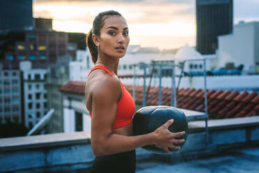 Fitness-Frau steht auf der Terrasse eines Gebäudes und trainiert mit einem Medizinball. Seitenansicht einer Frau in Fitnesskleidung, die auf dem Dach Übungen macht. - JLPSF19671