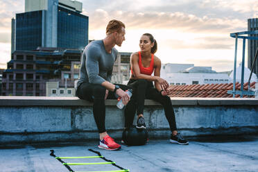 Man and woman in fitness clothes relaxing on the rooftop during workout. Fitness couple taking a break during workout sitting together on the fence of a terrace and talking. - JLPSF19672
