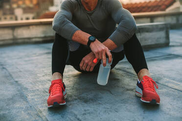 Fitness Mann sitzt auf dem Dach mit einer Wasserflasche nach dem Training. Cropped Schuss eines Mannes, der eine Pause entspannend sitzen auf der Terrasse eines Gebäudes nach dem Training. - JLPSF19678