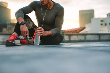 Male athlete relaxing after workout listening to music on earphones using mobile phone. Fitness man taking a break from workout sitting on rooftop holding cell phone and water bottle. - JLPSF19679