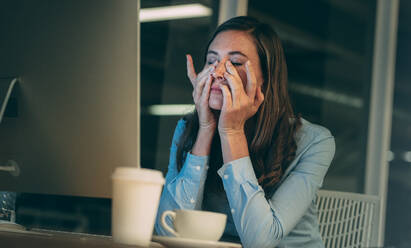 Unternehmerin schaut angespannt während der Arbeit spät im Büro. Geschäftsfrau sitzt müde vor dem Computer mit Händen auf dem Gesicht und geschlossenen Augen. - JLPSF19688