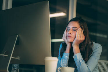 Geschäftsfrau sitzt vor dem Computer und sieht gestresst aus, wenn sie bis spät in die Nacht im Büro arbeitet. Frau sitzt im Büro mit Händen auf dem Gesicht und geschlossenen Augen und sieht müde aus. - JLPSF19689