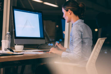 Woman doing graphic designing sitting at her desk in office late in the night. Woman using a digital writing pad to make designs on computer. - JLPSF19692