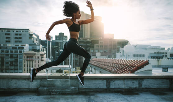 Weibliche Athletin läuft mit großen Schritten auf einem Dach am Morgen mit Sonne im Hintergrund. Frau in Fitnesskleidung trainiert auf der Terrasse eines Gebäudes. - JLPSF19700