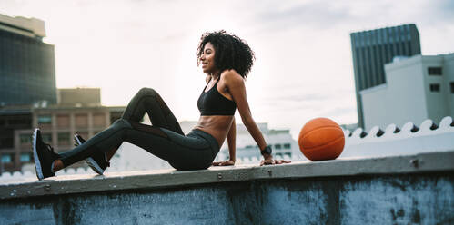 Frau in Fitnesskleidung sitzt entspannt auf einem Dachzaun und hat einen Basketball neben sich. Lächelnde Sportlerin bei morgendlichen Übungen auf der Terrasse eines Gebäudes. - JLPSF19711