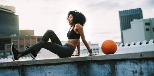 Woman in fitness wear sitting relaxed on rooftop fence with a basketball beside her. Smiling female athlete doing exercises in the morning on terrace of a building. - JLPSF19711