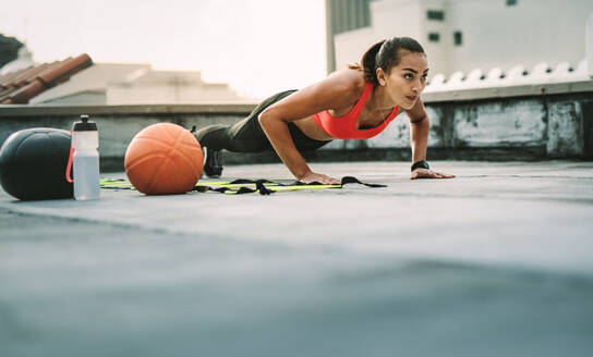 Frau Athleten tun Push-ups auf dem Dach mit Medizinball und Basketball an ihrer Seite. Frau in Fitness-Kleidung tun Workout auf der Terrasse. - JLPSF19714